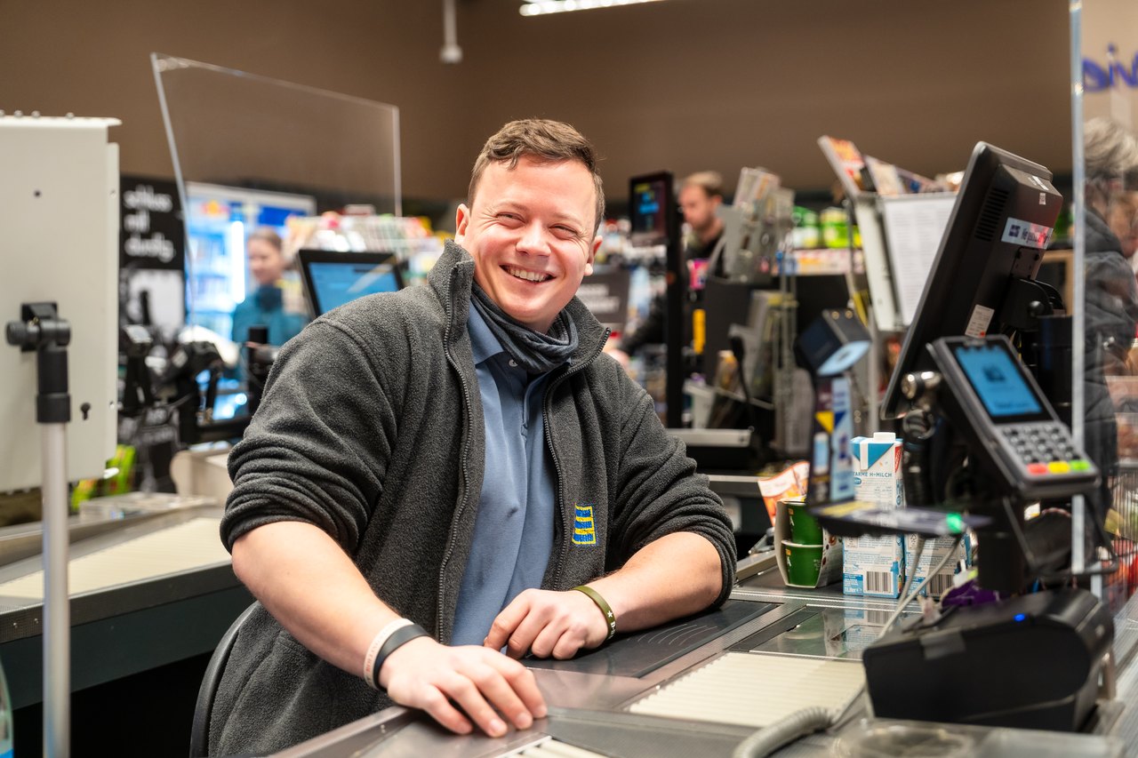 Ein lächelnder Edeka-Mitarbeiter steht hinter einer Supermarktkasse.