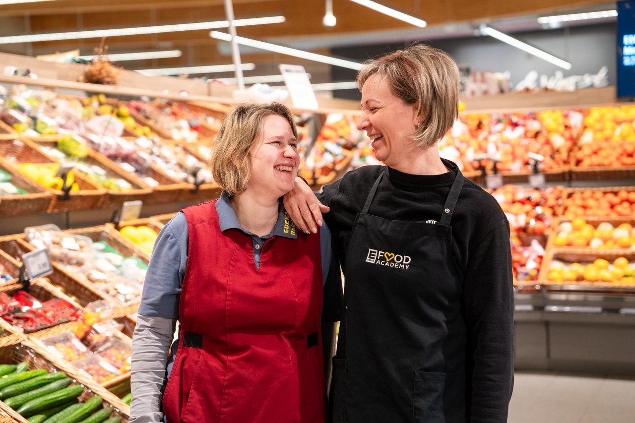 Zwei lächelnde Frauen in Schürzen stehen im Supermarkt vor Gemüseauslagen.