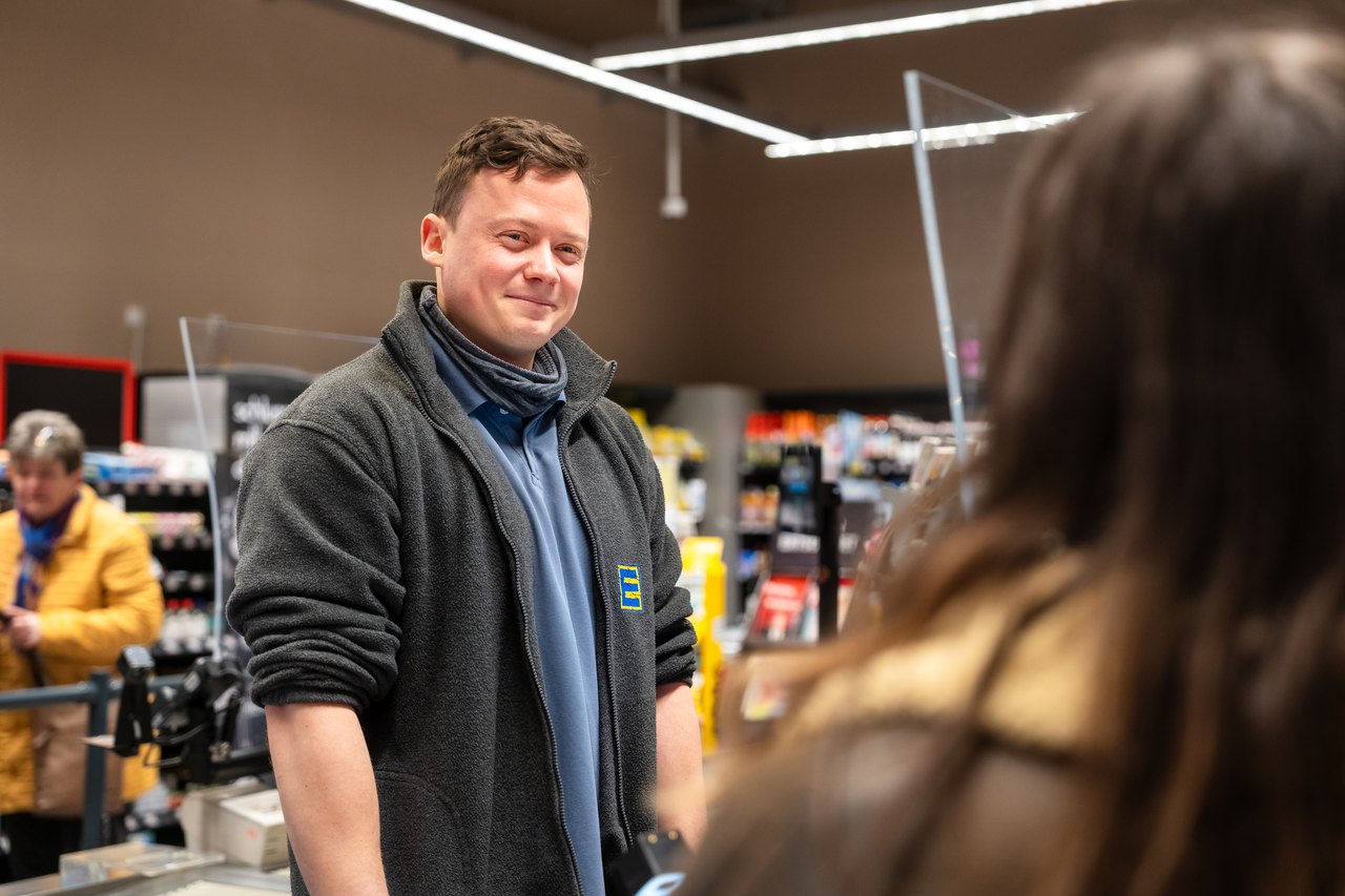 Ein lächelnder Edeka-Mitarbeiter in blauer und schwarzer Arbeitskleidung steht an einer Kasse.