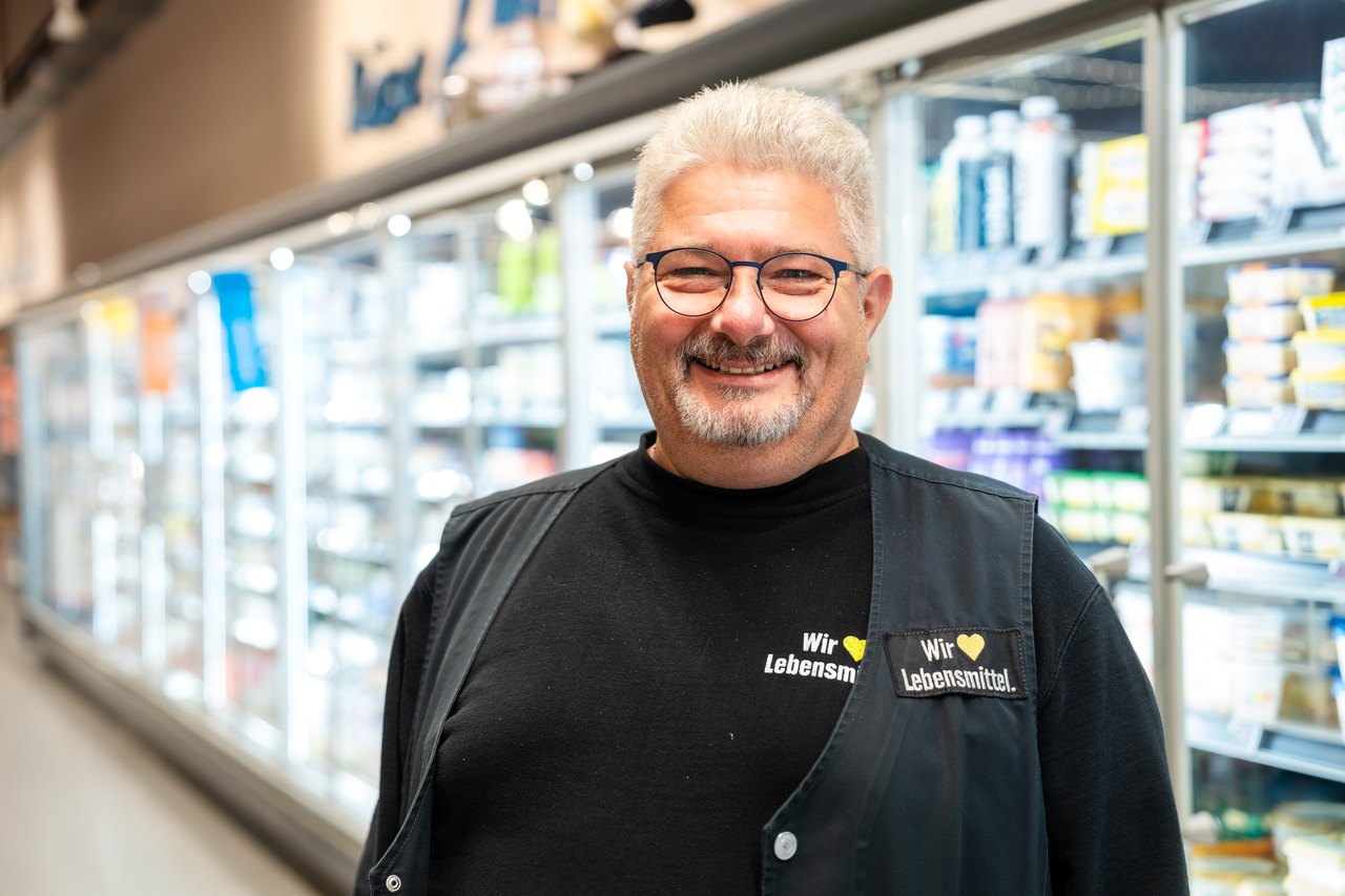 Lächelnder Mann mit Brille und grauem Haar in schwarzem T-Shirt mit gelben Logos vor Supermarkt-Kühlregalen.