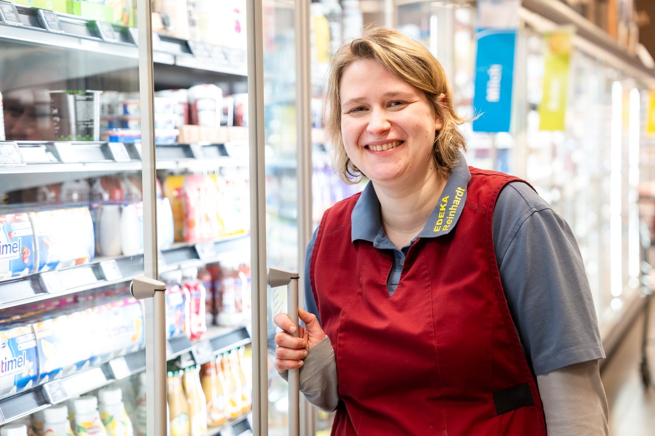 Lächelnde Frau im roten Kittel öffnet eine Glastür im Supermarkt-Kühlregal.