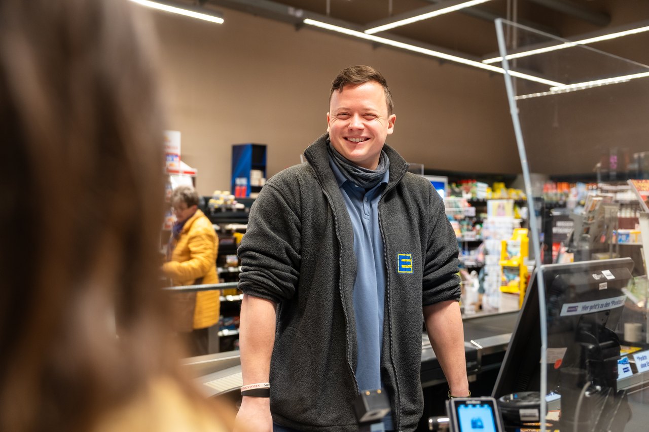 Ein lächelnder EDEKA-Kassierer in grauer Jacke steht an der Kasse und schaut eine Person an.