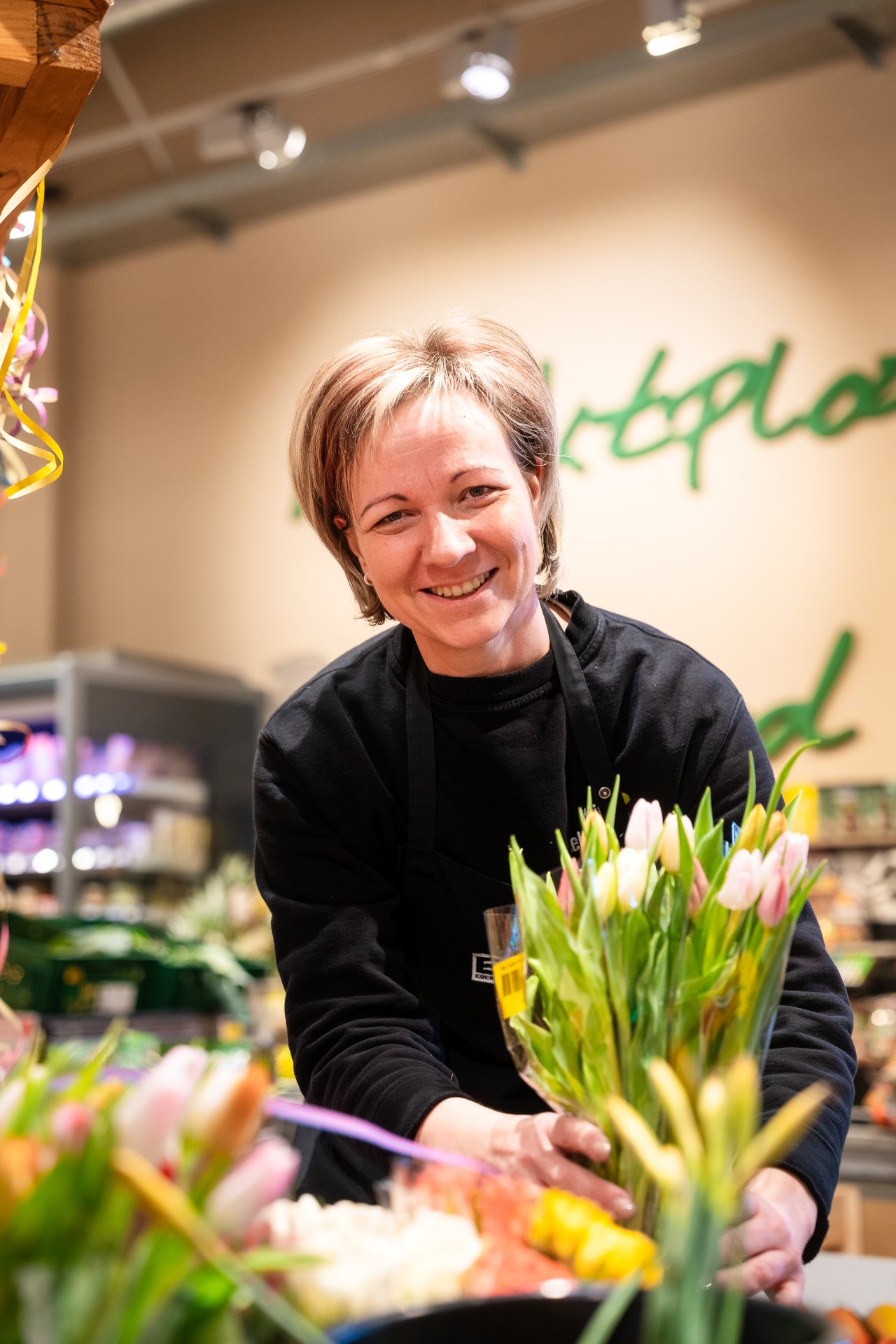 Eine lächelnde Frau in schwarzer Arbeitskleidung arrangiert einen Strauß rosa und weißer Tulpen.