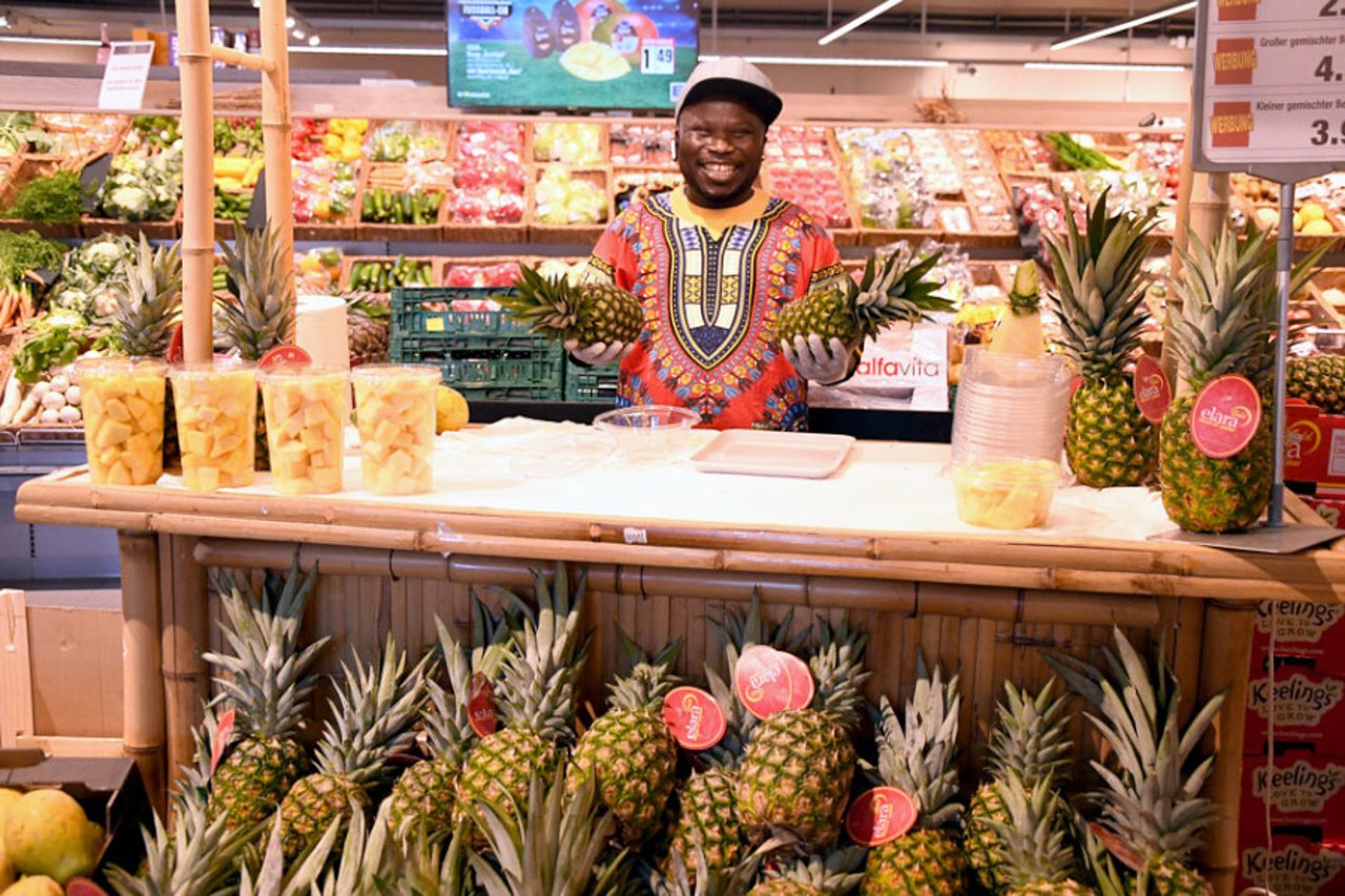 Ein Mann in einem bunten Hemd hält zwei Ananas hinter einem Bambusstand mit Ananas und Ananasstücken.