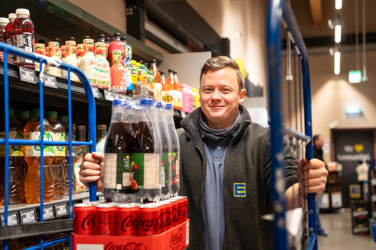 Junger Mitarbeiter im Edeka-Markt schiebt einen Rollwagen mit Getränken und lächelt in die Kamera.