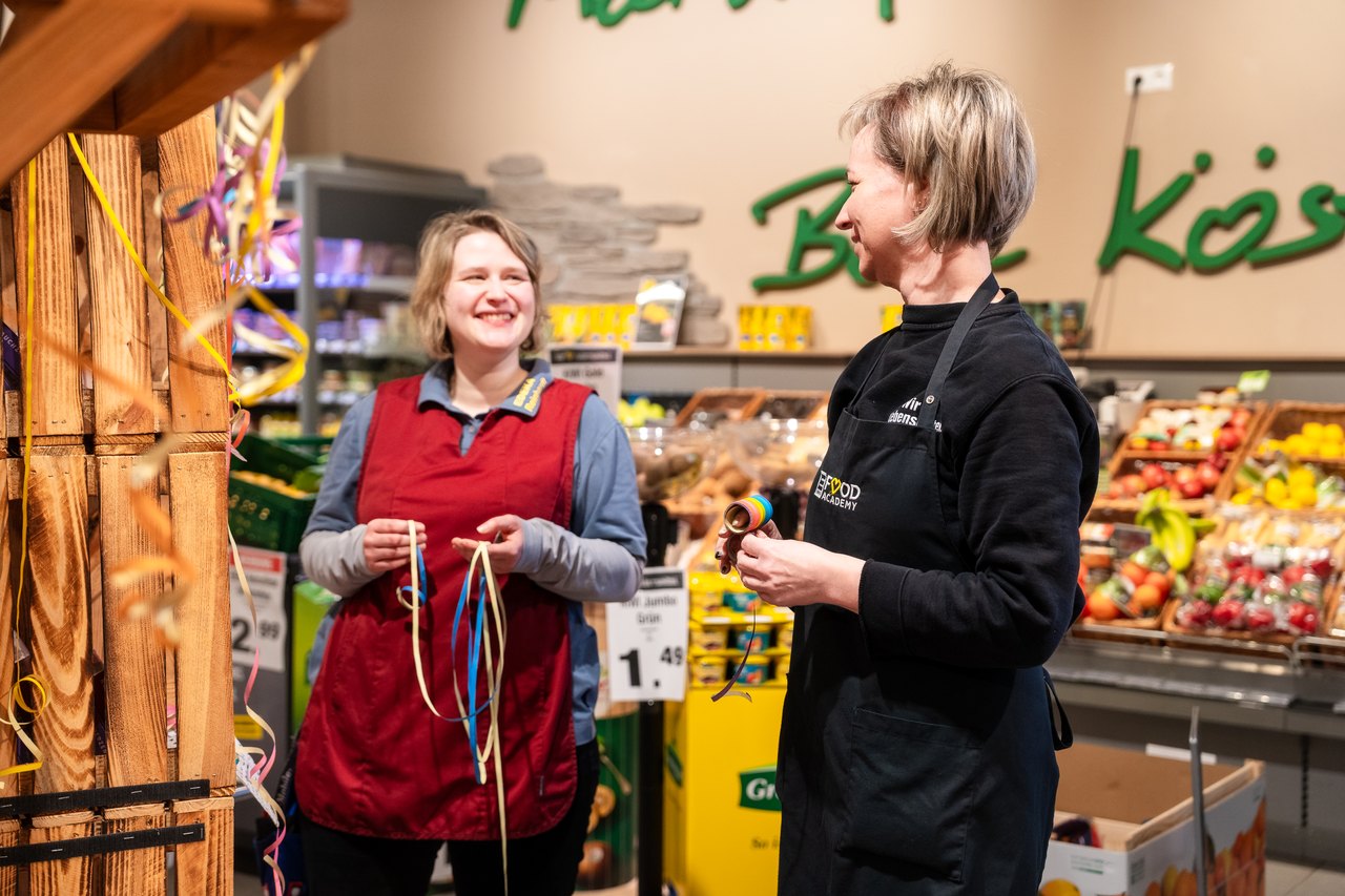 Zwei lächelnde Frauen in Schürzen in einem Supermarkt, eine hält Bänder, die andere spricht mit ihr.