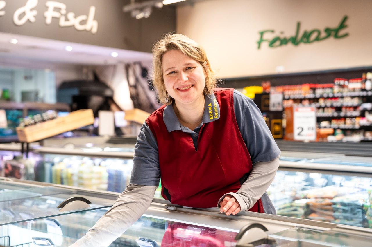 Lächelnde Frau in roter Weste und grauem Hemd beugt sich über eine Gefriertheke im Supermarkt.