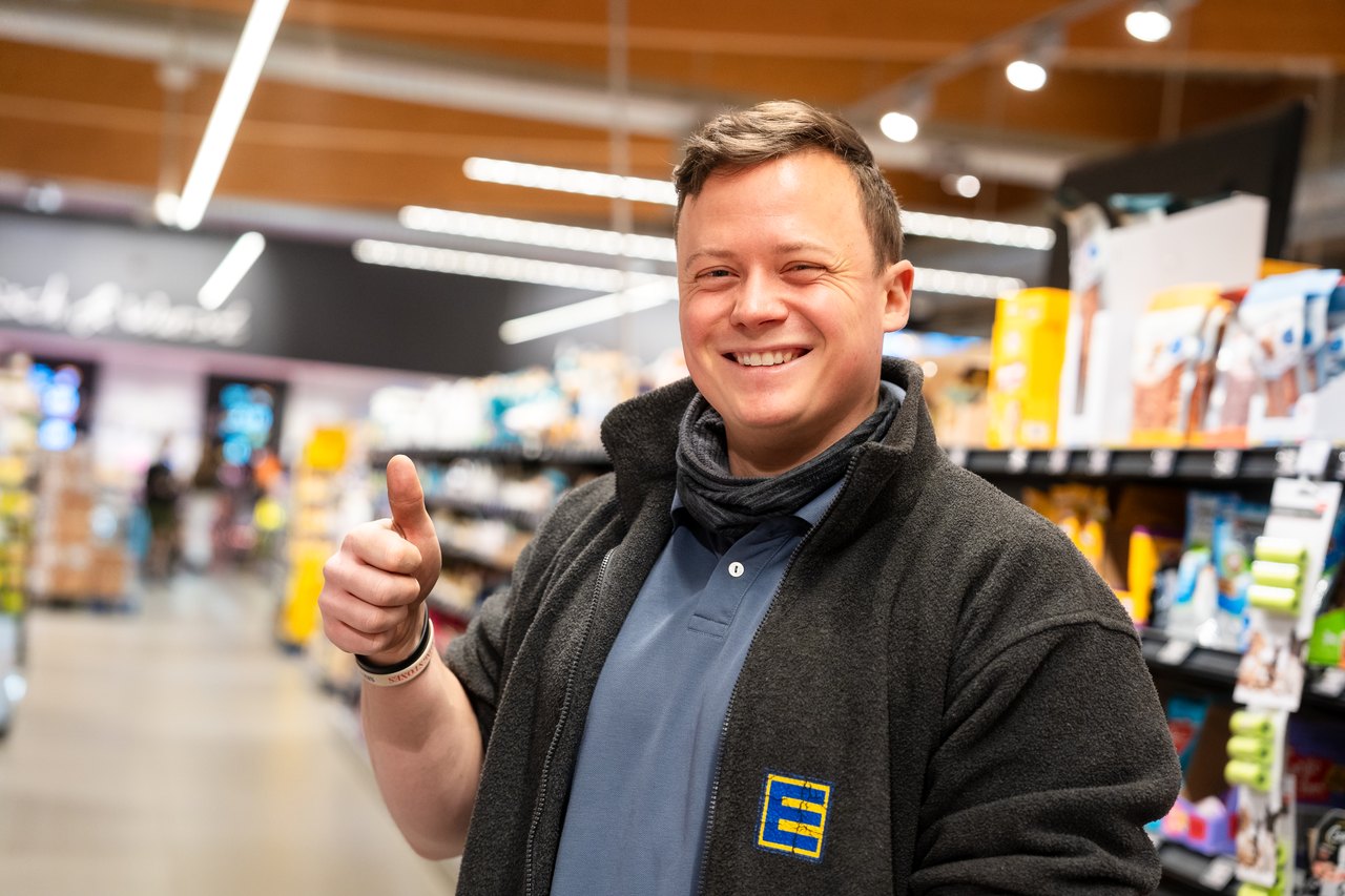 Ein lächelnder Mann in einer grauen Fleecejacke mit Edeka-Logo zeigt den Daumen nach oben in einem Supermarkt.