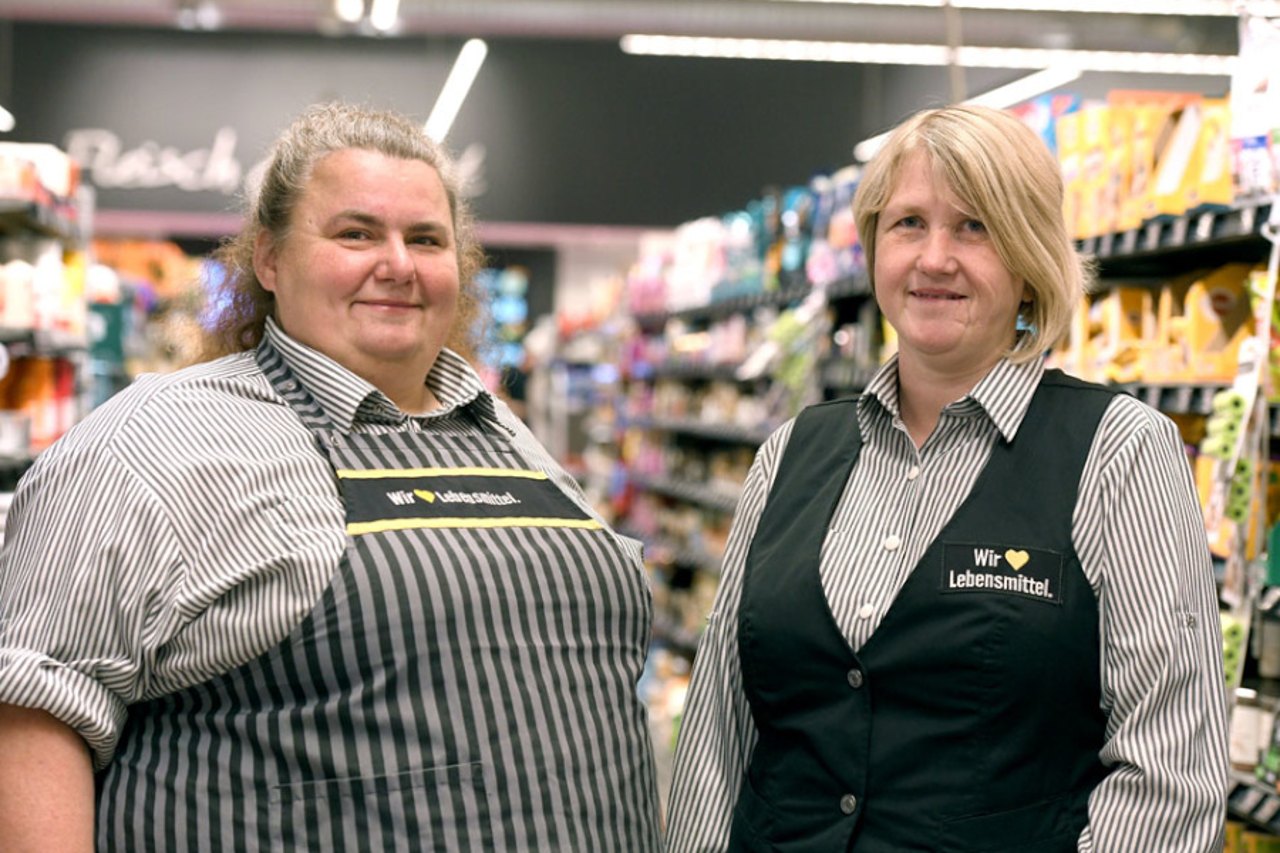 Zwei lächelnde Frauen in gestreiften Hemden und Schürzen/Westen mit „Wir Lebensmittel“ Logo in einem Supermarkt.